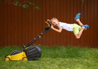 A blond boy is mowing the lawn in the garden with a yellow lawn mower. On a sunny summer day, a six-year-old boy mows the grass of a yellow lawn.