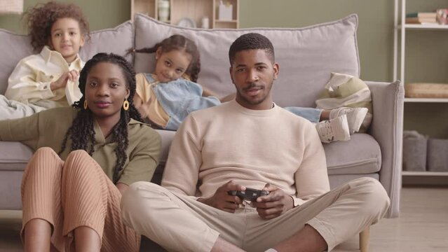 Slowmo of delighted young Black man using controller while playing video games at home while his beautiful wife and two pretty little daughters watching and cheering