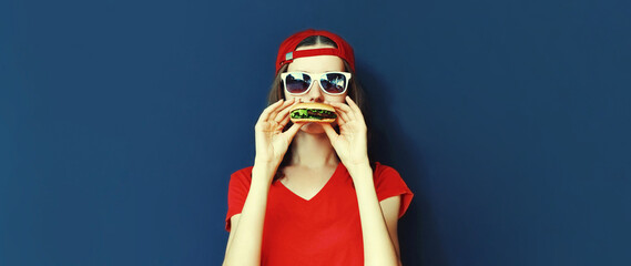 Portrait of stylish young woman eating tasty fast food burger wearing baseball cap, sunglasses on blue background