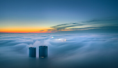 Skyscraper rooftop over the clouds at sunrise. Thick fog covers the Riga city, and warm sunlight over the clouds and church tower. - obrazy, fototapety, plakaty