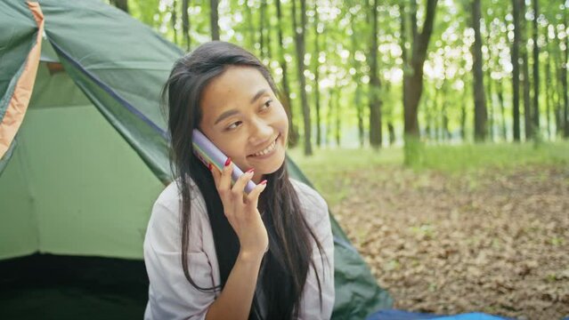 happy asian woman enjoy vacation, talking on video call, watching movie on phone, while relaxing with tent in forest city park. summer camping.