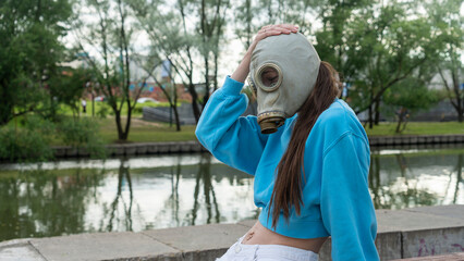 A girl in a gas mask sits on a bench on the river bank. Alienation in the city. Fashion, ecology...