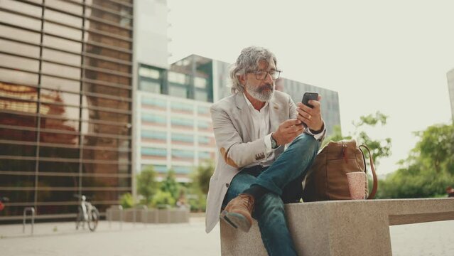 Mature businessman with beard in eyeglasses wearing gray jacket is using cell phone. Middle aged manager scrolling information on his smartphone while sitting outside the office
