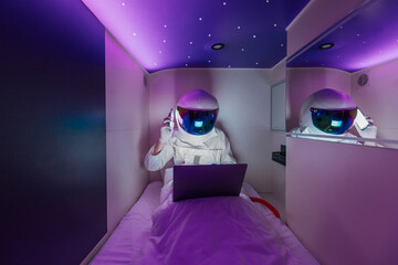 Spaceman work on a laptop and talking to a smartphone in capsule hotel sleeping module