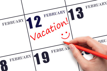 A hand writing a VACATION text and drawing a smiling face on a calendar date 12 February. Vacation planning concept.