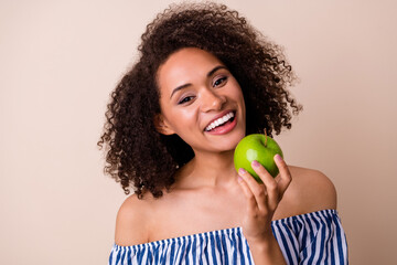 Photo of sweet pretty girl dressed off shoulders clothes eating green apple smiling isolated beige...