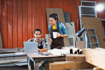 Young Asian couple carpenters man and woman discussing about design of products on laptop in woodcraft carpentry workshop.