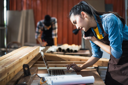 Young Asian female carpenter using laptop computer while talking to customer on the phone in woodcraft carpentry workshop.