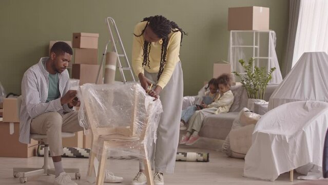 Slowmo Of Married African American Couple With Two Little Daughters Unwrapping Furniture From Plastic After Moving In New Apartment