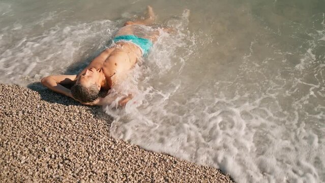 handsome athletic man in blue swim trunks relaxing by lying on pebble beach close to turquoise water of mediterranean sea in Oludeniz Beach in Aegean Sea after swimming and sunbathing. summertime wibe