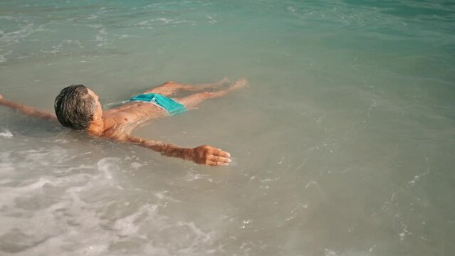 slow motion handsome man in blue swim trunks relaxing by lying in shallow water in turquoise mediterranean sea in Oludeniz Beach in Aegean Sea after swimming and sunbathing. summertime mood and relax