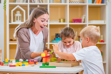 Kindergarten teacher playing together with children in the colorful preschool classroom. Mother playing with children. - obrazy, fototapety, plakaty