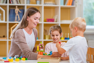 Fototapeta premium Kindergarten teacher playing together with children in the colorful preschool classroom. Mother playing with children.