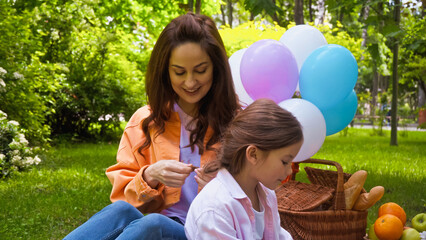 happy mother braiding hair of daughter near balloons in park.