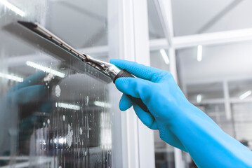 A man wearing light blue rubber gloves cleans the surface of an interior office window with a glass...