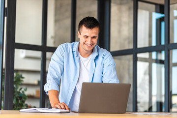 Fototapeta premium Confident entrepreneur standing in the office near the workplace and working using a laptop. Happy male young businessman with computer, looking at the screen, smiles friendly. Distant work