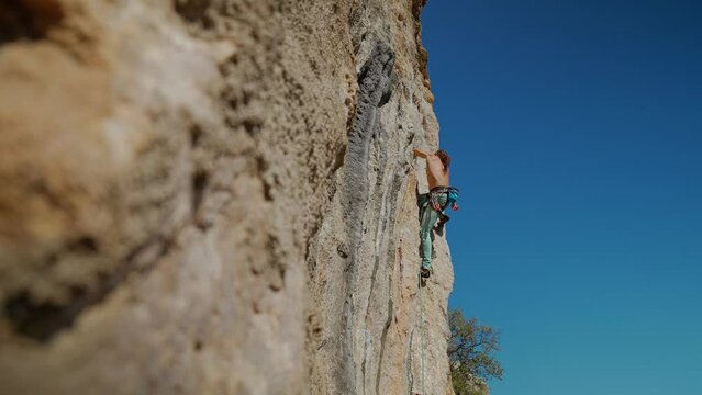 bottom view of muscular strong man rock climber climbs on vertical cliff on rock wall on blue sky background. man puts quickdraw and clips a rope to make belay and be in safe