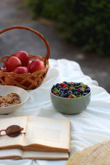 Picninc blanket with straw bag, bowl of strawberries and blueberries, bowl of chocolate chip cookies, books, sunglasses and basket of apples in the garden. Selective focus.