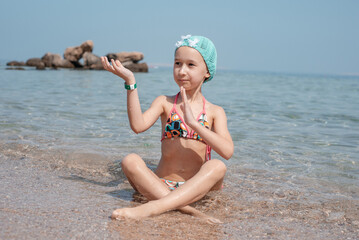 A little girl in a colored swimsuit and a blue cap is doing yoga on the seashore. Happy girl by the sea. Summer holidays, travel concept. Meditation by the sea.