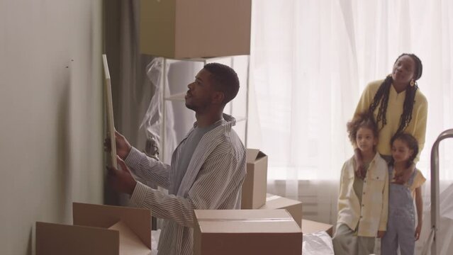Slowmo of Black man hanging picture on pastel olive green wall while settling in new apartment after moving in with beautiful wife and two pretty little daughters