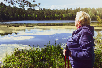 Elderly woman with nordic sticks stand by blooming lake in spring outdoors in forest. Kurtuvenai...
