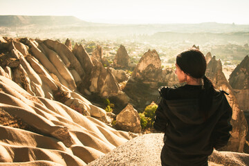 Thoughtful Female person stand look over dramatic valley on hazy morning sunrise with fairy chimneys background. Solo exploration in Turkey. Cinematic Travel destination-Cappadocia - obrazy, fototapety, plakaty