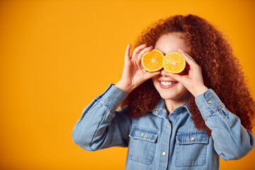 Studio Portrait Of Girl Holding Two Orange Halves in Front Of Eyes Against Yellow Background