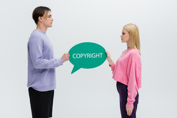 Side view of young couple in sweatshirts holding speech bubble with copyright lettering isolated on grey.