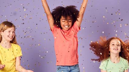 Studio Shot Of Children With Glitter Jumping In The Air With Outstretched Arms On Purple Background