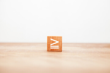 ≧の記号。大なりイコール。不等号。同じか大きい。木製ブロックに書かれている。白い文字。木製テーブルの背景。