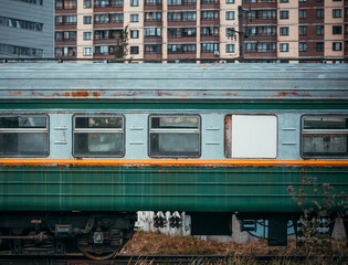 An old passenger railway car with peeling paint in the city on the background of houses and buildings.  An empty carriage on the railway.