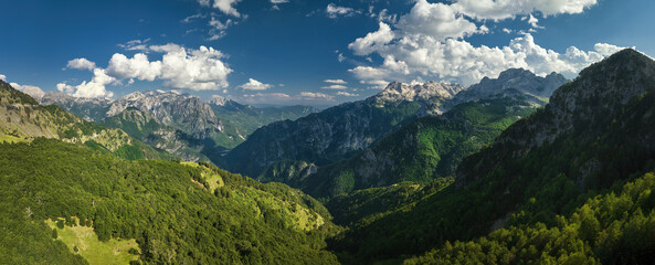 Naklejka na ściany i meble A panoramic, aerial view of the monumental Albanian Alps. Hiking trails, dark blue sky with clouds, steep rocks, green valley, remnants of snow, summer. Theth National Park, Albanian Alps, Albania.