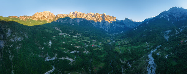 A panoramic, aerial view of the Theth valley during sunset. Hiking paths, green valleys, blue sky with clouds, steep rocks, remnants of snow, summer. Theth national park, Albanian Alps, Albania.
