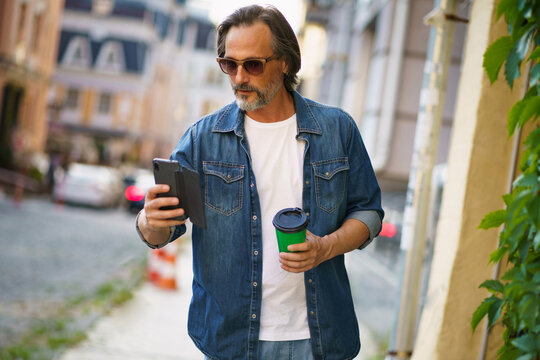 Mature man read text or email walking off work or back to office holding phone and take away paper cup with hot coffee wearing sunglasses with at old town streets. Business on the go concept