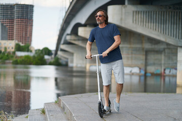 Riding scooter handsome stylish middle aged man with grey beard stand under town bridge over river with urban city on background after work outdoors. Travel, lifestyle concept