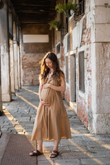 Pregnant woman in summer dress touching belly on urban street in Venice.