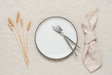 Autumn table setting. A white plate, cutlery, a beige linen napkin and ripe ears of wheat on a...