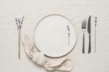 Table setting. Empty beige plate, cutlery, linen napkin and dried lavender flowers on a beige linen tablecloth. Top view, flat lay.
