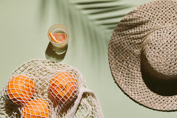 Summer flat lay with straw hat, glass of water and orange fruit in eco shopping bag. Green background with palm leaf shadow, sun and sunlight. Vacation, holiday, summer creative minimal concept. © magic_cinema