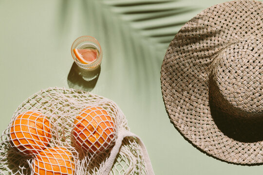 Summer flat lay with straw hat, glass of water and orange fruit in eco shopping bag. Green background with palm leaf shadow, sun and sunlight. Vacation, holiday, summer creative minimal concept.