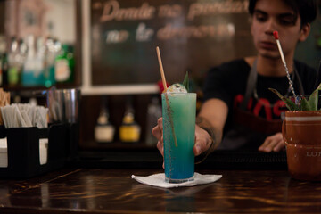 Bartender of a nightclub at the bar counter serving a blue-colored drink for a customer. Blue drink served by a waiter. A colorful alcoholic drink with ice cubes.