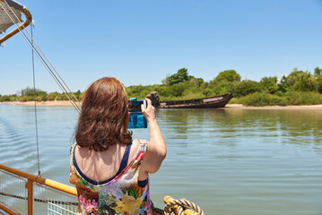 Naklejka premium Back view of an hispanic woman taking pictures with her phone from a tourist boat sailing on the Uruguay River in Colon, Entre Rios, Argentina. Concepts: summer tourism, enjoying the outdoors.