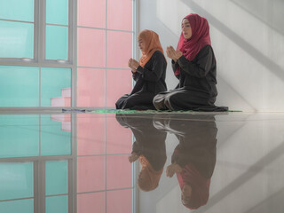 Two pretty Muslim women in hijab with colorful headscarf sitting praying on carpet. Moslem pray, praying hands to Allah of Muslim ladies. The shiny floor showing reflection.