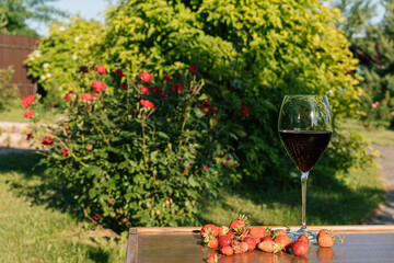 A glass of red wine and strawberries against outdoor garden background.