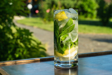 Cold iced mineral water with mint leaves and lemon pieces in tall glass on a table on outdoor background.