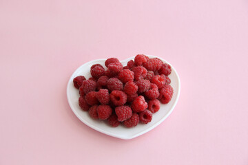 large ripe raspberries in a white plate in the shape of a heart on a pink background