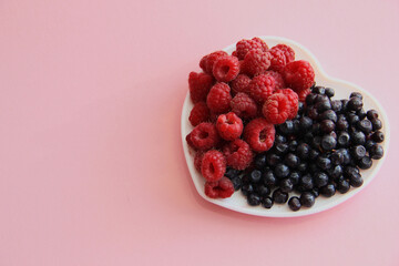 ripe raspberries and blueberries on a white plate in the shape of a heart on a pink background