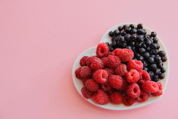 ripe raspberries and blueberries on a white plate in the shape of a heart on a light background close-up