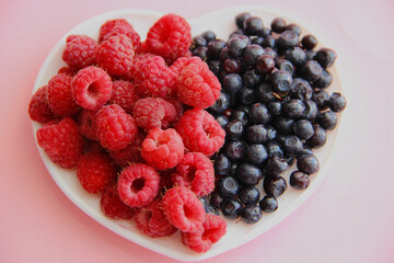 ripe raspberries and blueberries on a white plate in the shape of a heart on a pink background