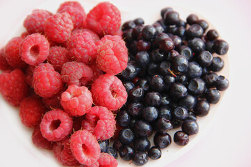 Large raspberries and blueberries on a white plate in the form of a heart on a pink background